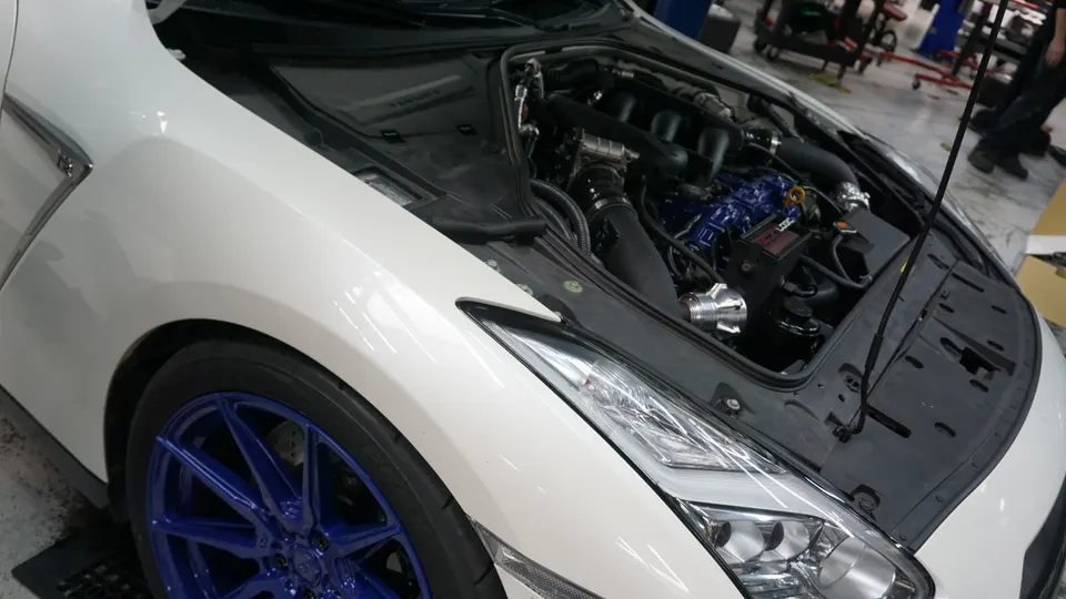 A white Nissan GT-R with its hood open, revealing a modified engine bay with blue components and an aftermarket intake in a shop.