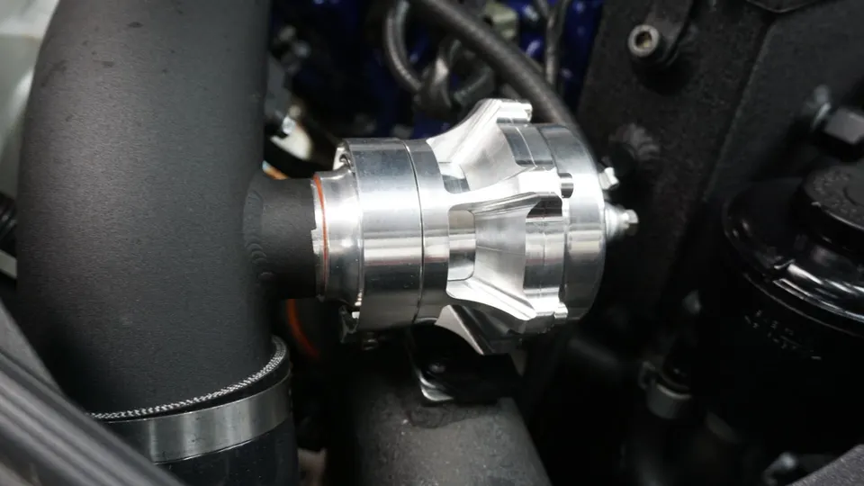 Close-up of a polished blow-off valve installed in an engine bay with black boost piping, part of a turbo system.