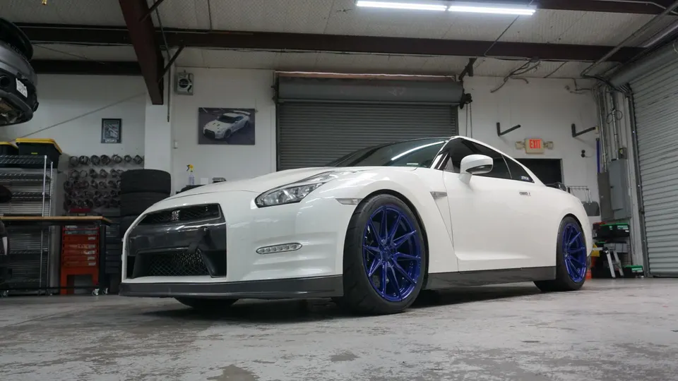 A white Nissan GT-R with blue aftermarket wheels parked inside a performance tuning shop.
