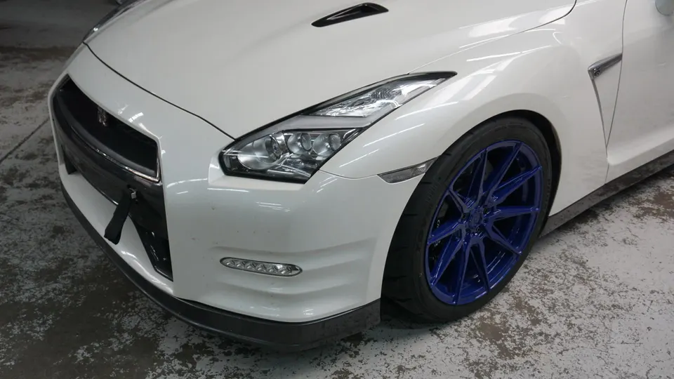 White Nissan GT-R with custom blue multi-spoke wheels, showing the front bumper, splitter, and LED headlights.