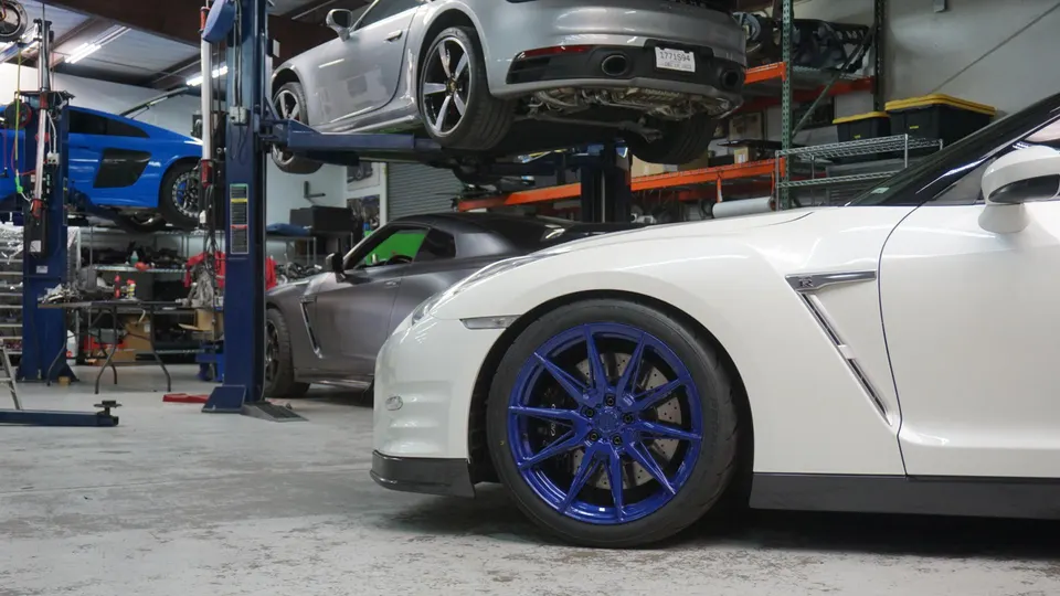 A white Nissan GT-R with blue wheels in a performance tuning shop, with a silver Porsche 911 and a blue Audi R8 on lifts.