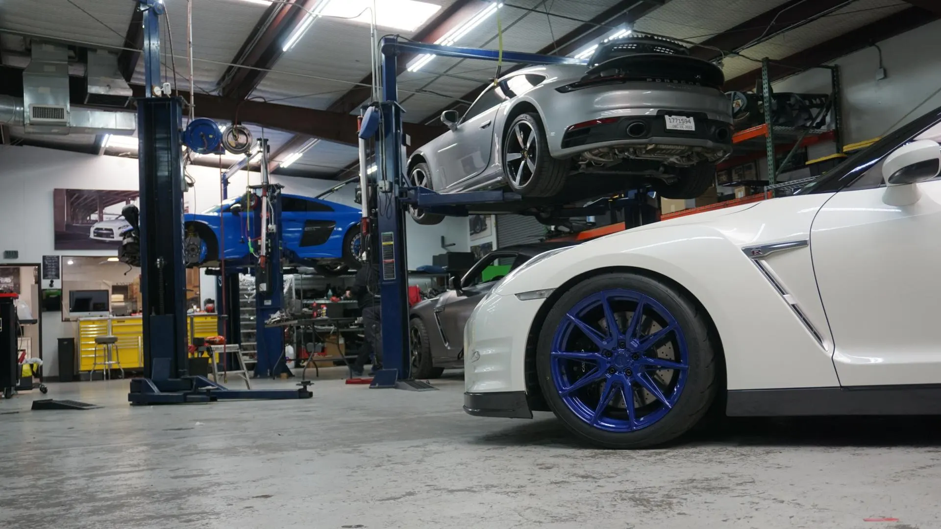 A white Nissan GT-R with blue wheels in the foreground of a car shop, with a silver Porsche 911 and a blue Audi R8 on lifts.