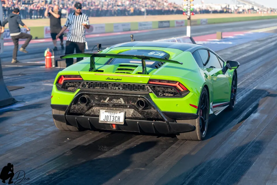 Rear view of a green Lamborghini Huracan with a large rear wing and diffuser on a drag strip, with a starting tree in the background.