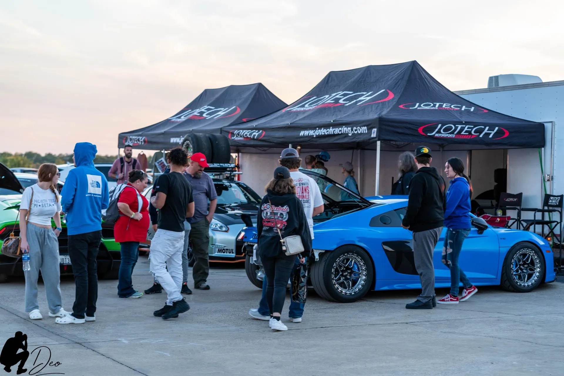 People gather around a blue Audi R8 and a silver Nissan GT-R under Jotech Motorsports tents at a car event.