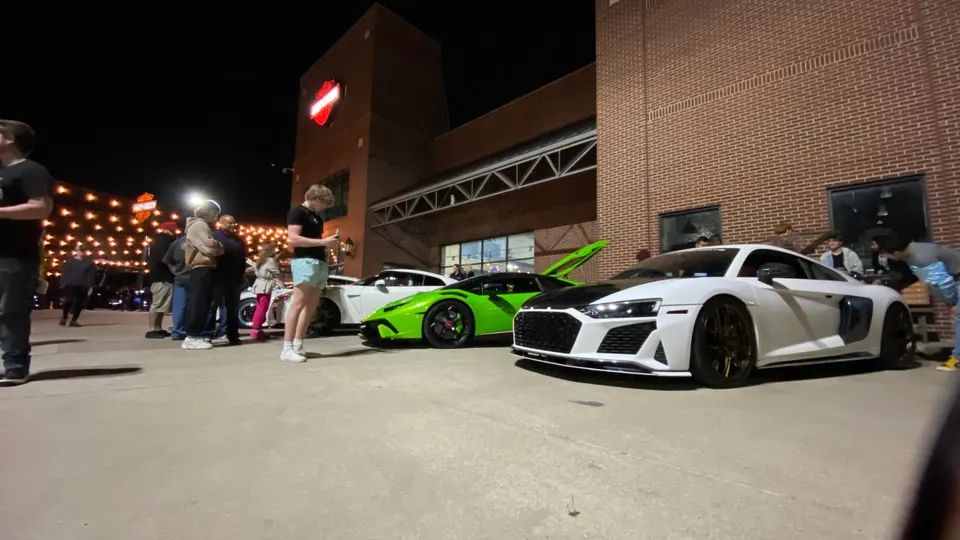Audi R8 and Lamborghini Huracan supercars displayed at a night car meet with a crowd of enthusiasts.