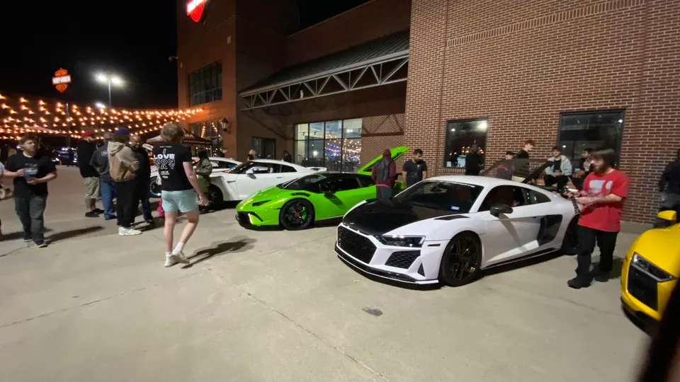 Supercars including a green Lamborghini Huracan and white Audi R8 at a night car meet with a crowd of enthusiasts.