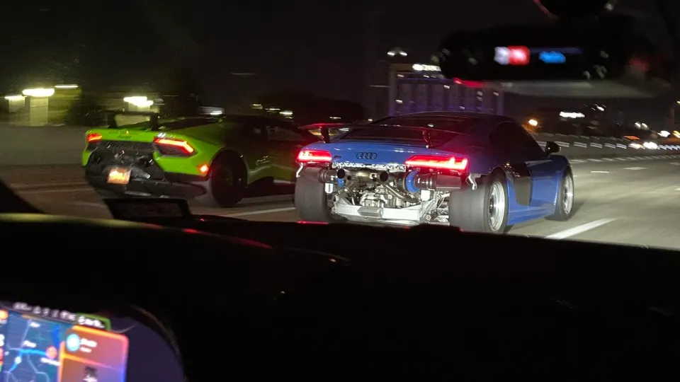 Audi R8 with exposed twin turbos and drag tires next to a green Lamborghini Huracan on a road at night.