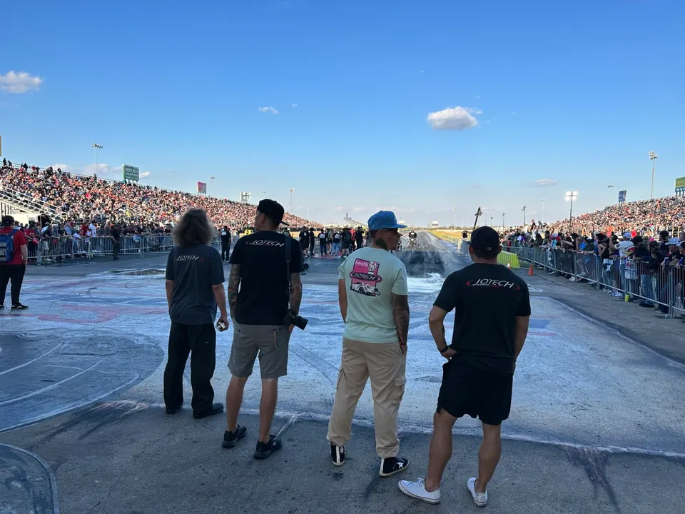 Four people wearing Jotech shirts stand on a drag strip, looking down the track with large crowds in the grandstands.