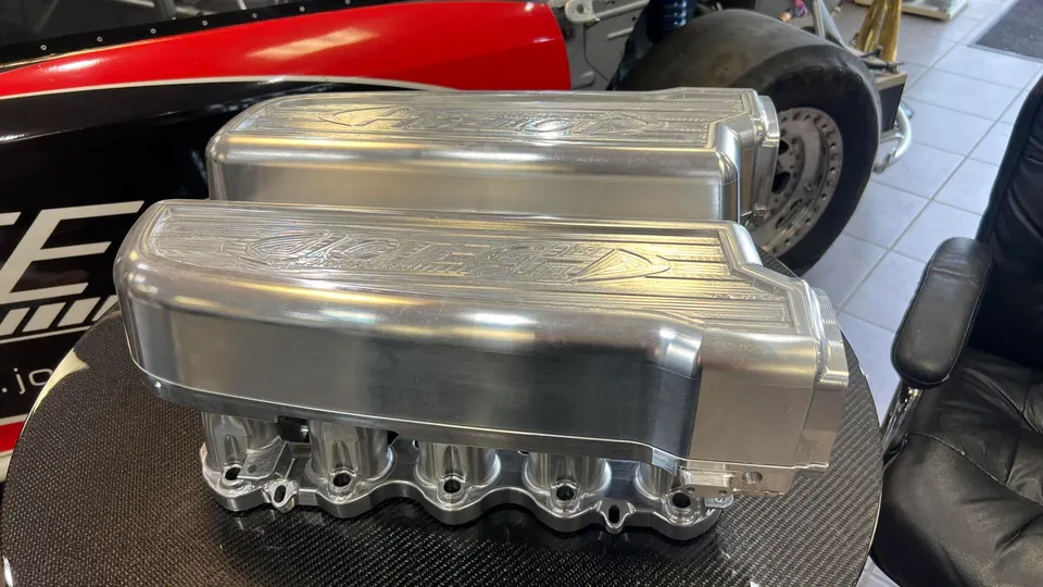 Two polished billet intake manifolds on a carbon fiber table in a Jotech Motorsports shop, with a race car in the background.