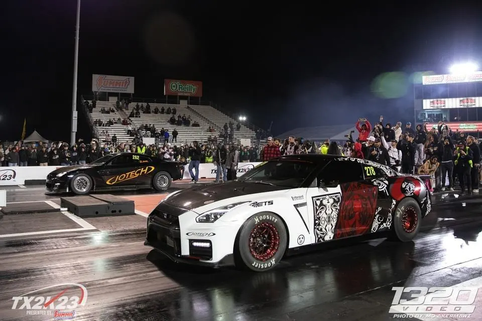 Two Jotech Motorsports Nissan GT-Rs on a drag strip at night, with spectators in the stands.