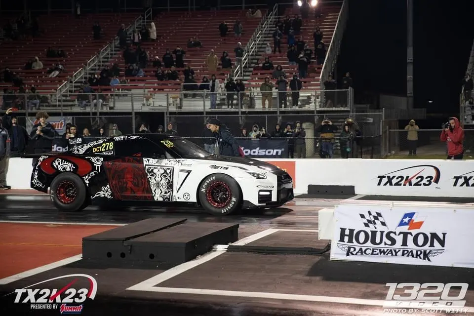 A Nissan GT-R with a custom black and red wrap at the starting line of a drag strip during TX2K23.