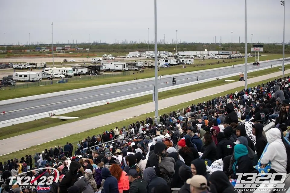 Spectators fill the bleachers at a drag racing event, with the track and RVs in the background.