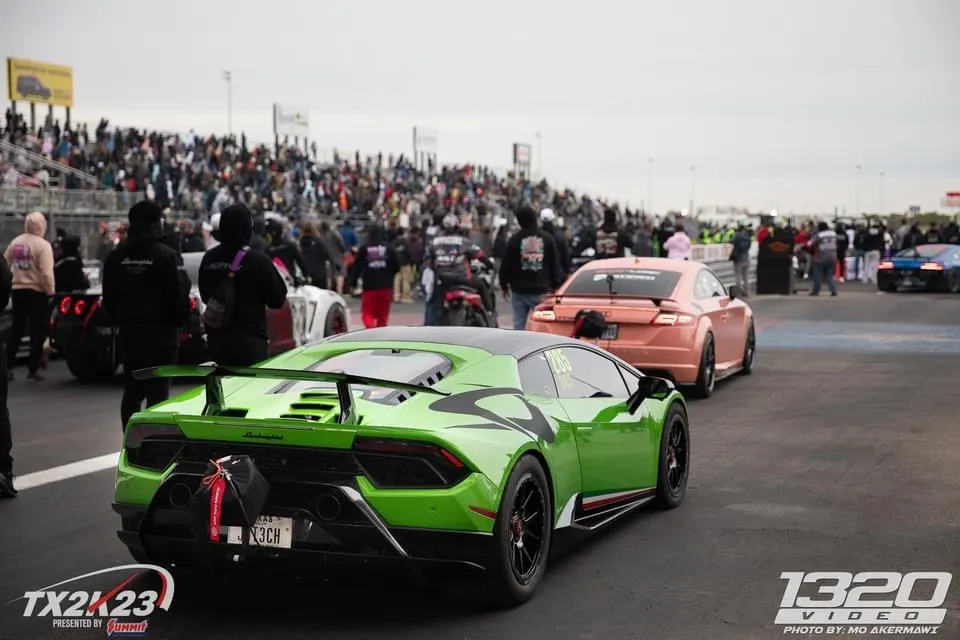 A green Lamborghini Huracan and a pink Audi TT in the staging lanes at a drag strip event with spectators.