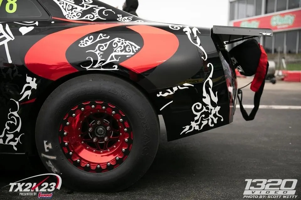 A black and red custom-liveried drag car with a large rear wing, parachute, and red beadlock drag wheels on a track.