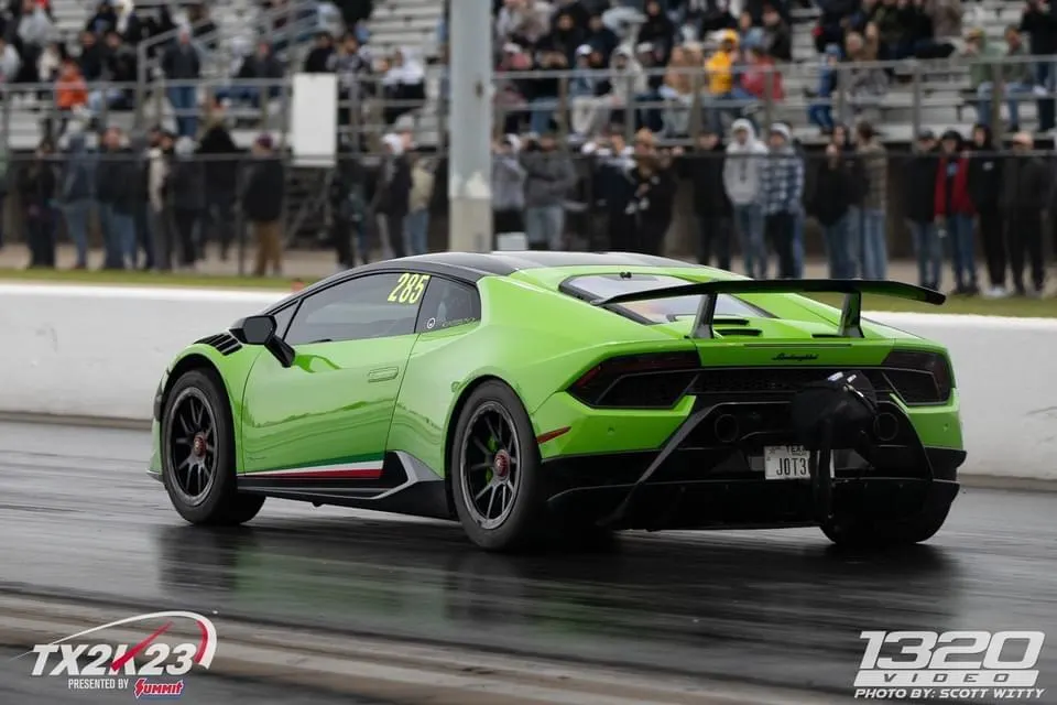 A bright green Lamborghini Huracan with a large rear wing and a parachute on a drag strip, with spectators in the background.