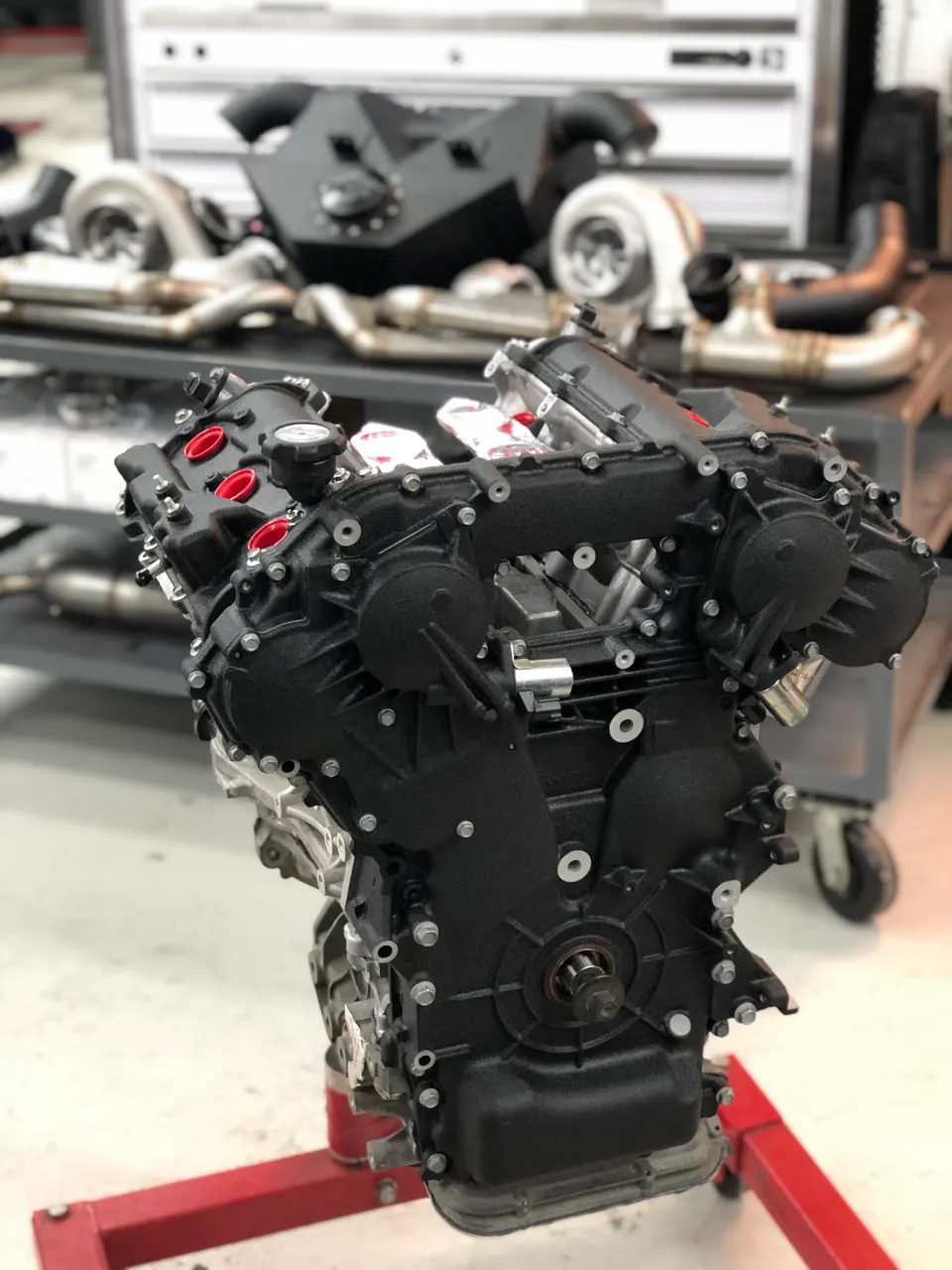 A built performance engine on a stand in a tuning shop with turbochargers and exhaust components in the background.