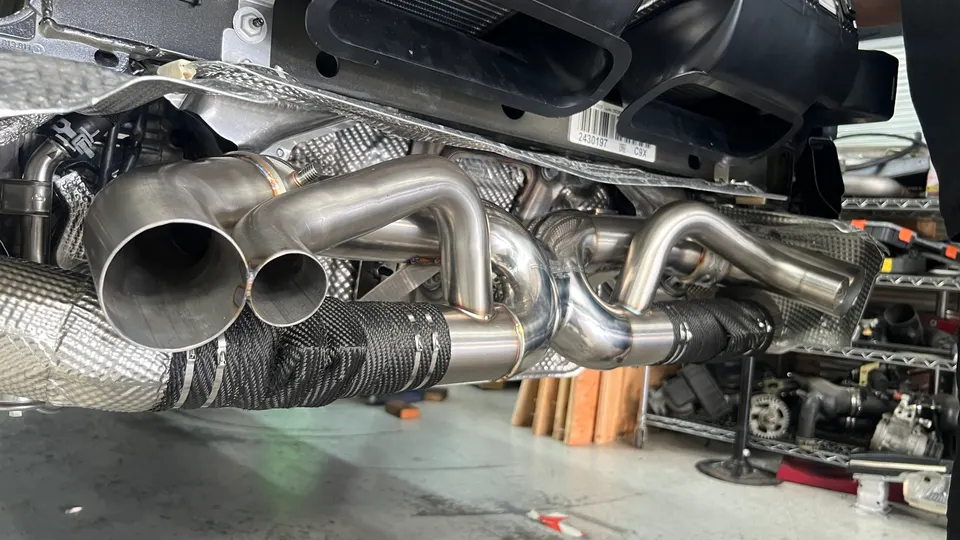 Close-up of a custom stainless steel exhaust system with heat wrap being installed on a performance car in a shop.