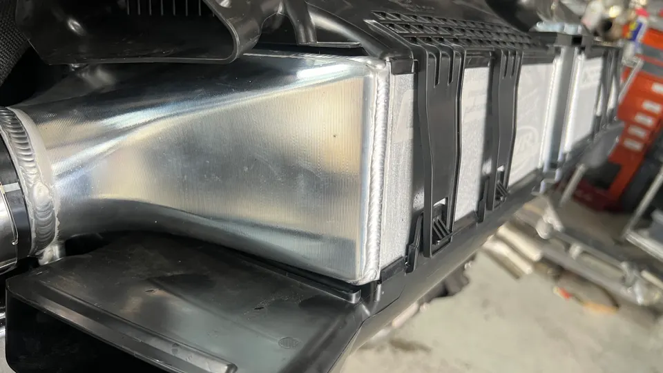 Close-up of a performance aluminum intercooler or charge pipe with visible welds in an engine bay.