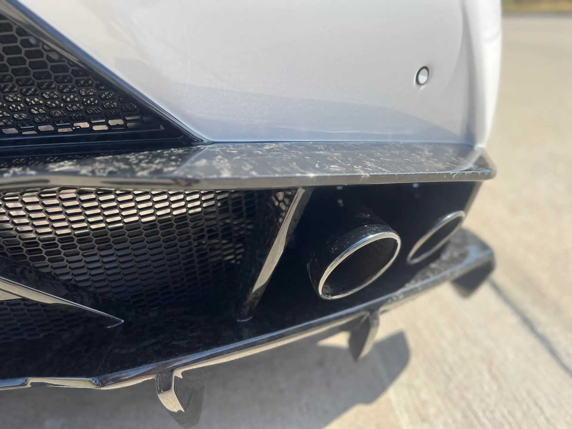 Close-up of a white car's rear bumper featuring a carbon fiber diffuser and dual exhaust tips.