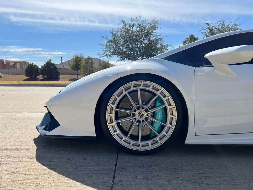 A white Lamborghini supercar with custom multi-spoke wheels and teal brake calipers, parked outdoors on a sunny day.
