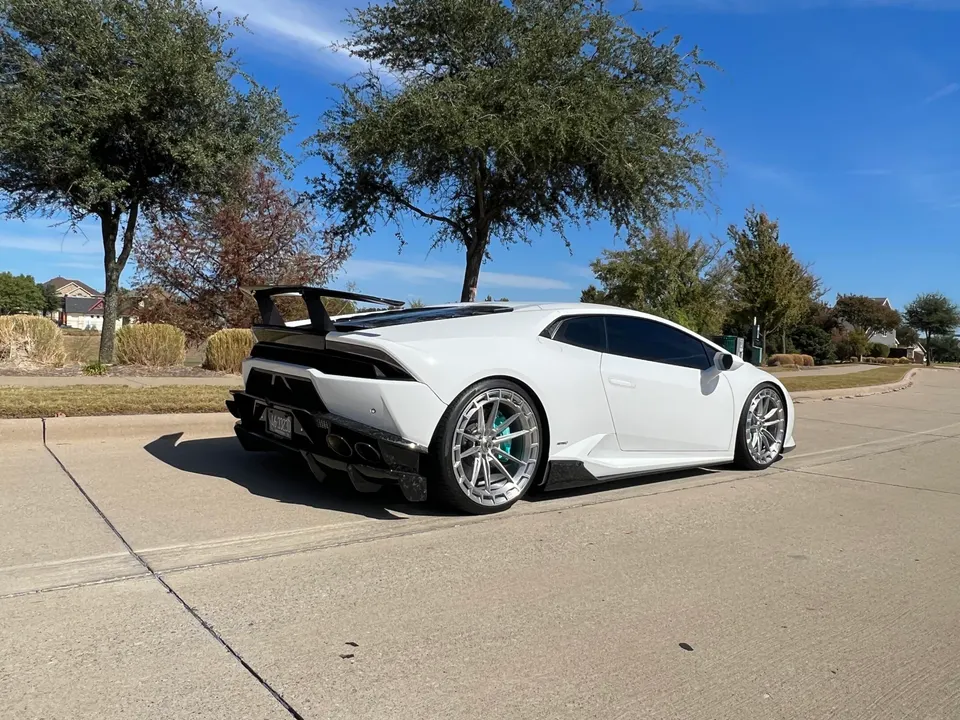 A white Lamborghini Huracan with aftermarket wheels and a carbon fiber rear wing parked on a residential street.