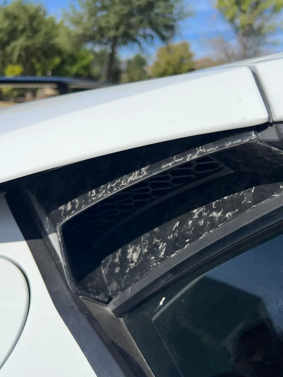 Close-up of a white supercar's side vent with forged carbon fiber trim, showcasing exterior aero detail.