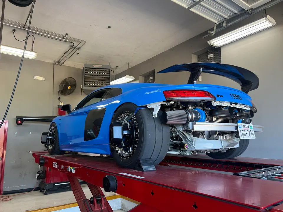 A blue Audi R8 with its rear bumper removed, revealing a twin-turbo setup and exhaust, on a shop lift for alignment.