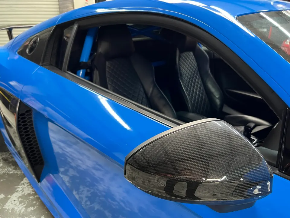 Close-up of a blue Audi R8 featuring a carbon fiber side mirror and a visible roll cage in the interior.