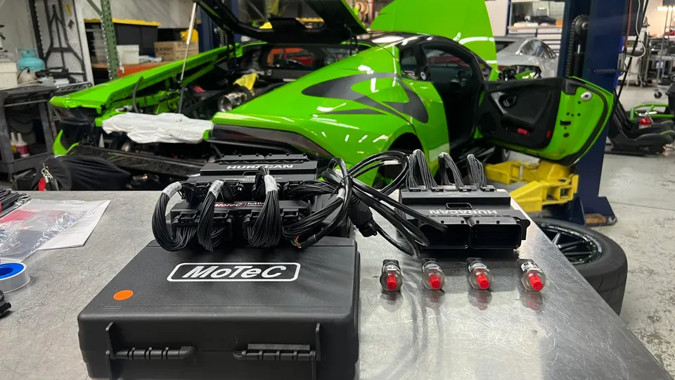 MoTeC ECU and wiring harnesses on a workbench in front of a green Lamborghini Huracan undergoing tuning at a performance shop.