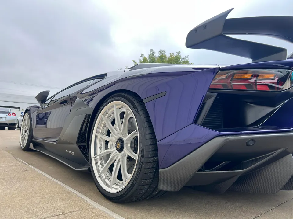 Purple widebody Lamborghini Aventador with custom wheels, large rear wing, and carbon fiber diffuser parked outdoors.
