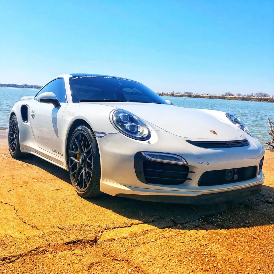 A white Porsche 911 Turbo S parked on a dirt bank next to a lake under a clear blue sky.