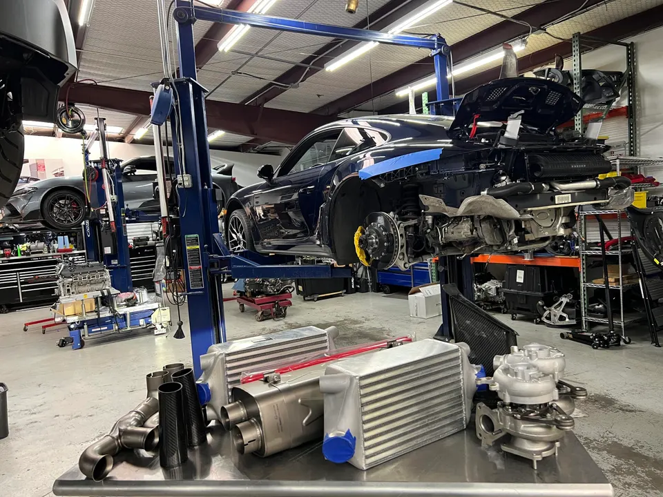 A Porsche 911 on a lift in a performance shop with turbochargers, intercoolers, and exhaust parts on a table.