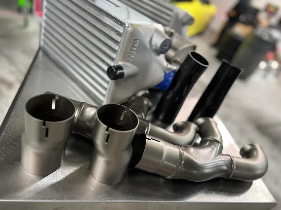 Performance car parts including an Alpha intercooler and titanium exhaust pipes laid out on a metal surface in a shop.