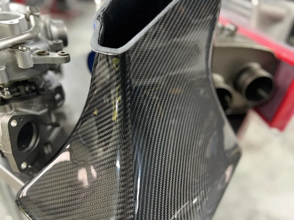 Close-up of a carbon fiber intake system for a performance car, with other engine components in the background.