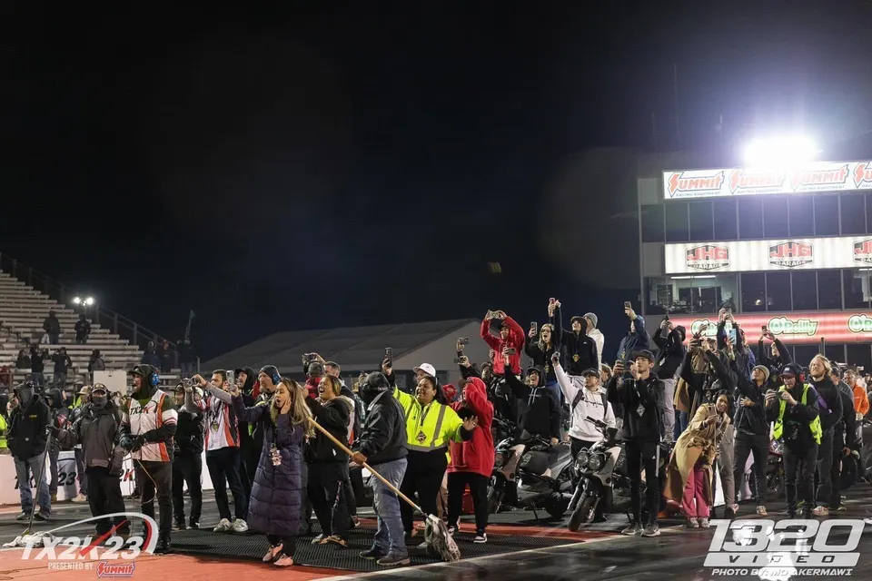A large crowd of spectators at a night drag racing event, many holding up phones to capture the action.