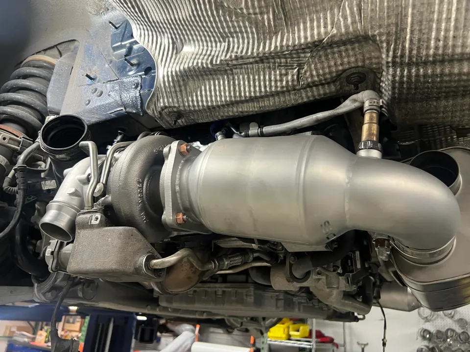 Underside view of a car showing a turbocharger and a new downpipe with catalytic converter being installed.