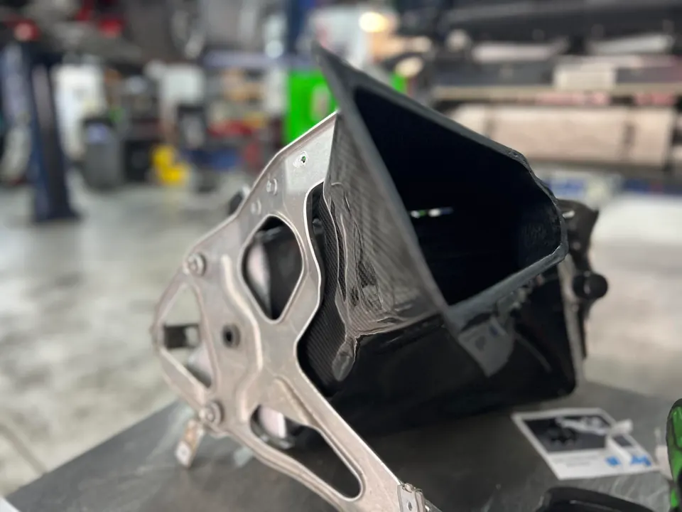 Carbon fiber air intake duct with a metal bracket on a workbench in a performance tuning shop.