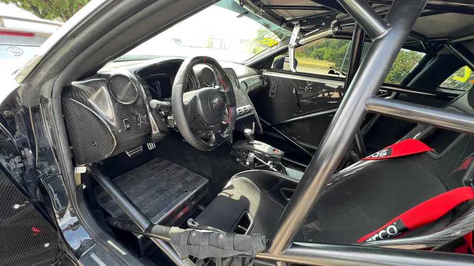 Interior view of a modified Nissan GT-R with a full roll cage, carbon fiber trim, racing seat, and Sparco harness.