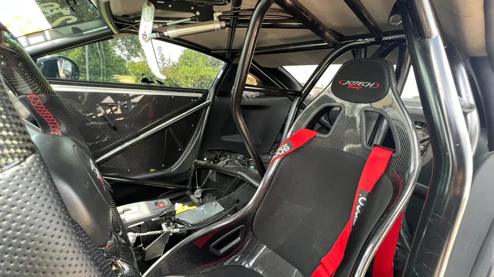 Interior view of a modified performance car featuring a Jotech Racing carbon fiber racing seat, red Sparco harness, and a black roll cage.