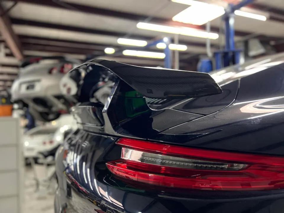 A dark blue Porsche 911 with a carbon fiber rear wing and LED taillights, parked in a performance tuning shop.