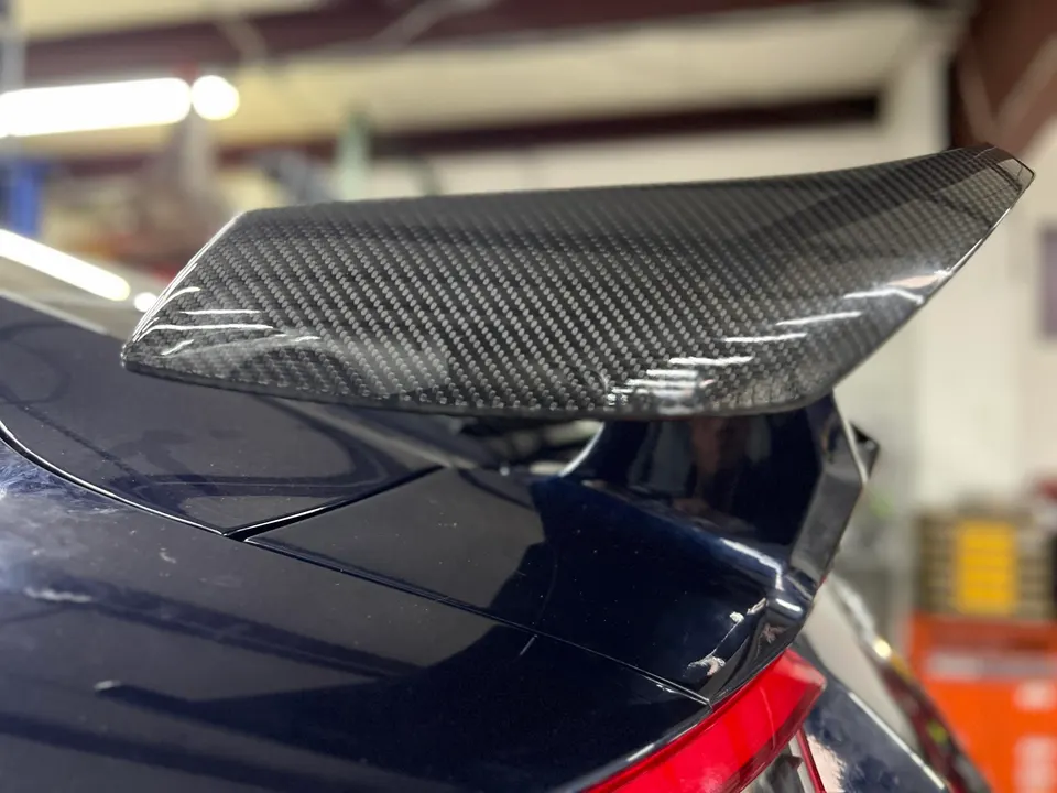 Close-up of a carbon fiber spoiler on the rear of a dark blue car, showcasing the weave pattern.