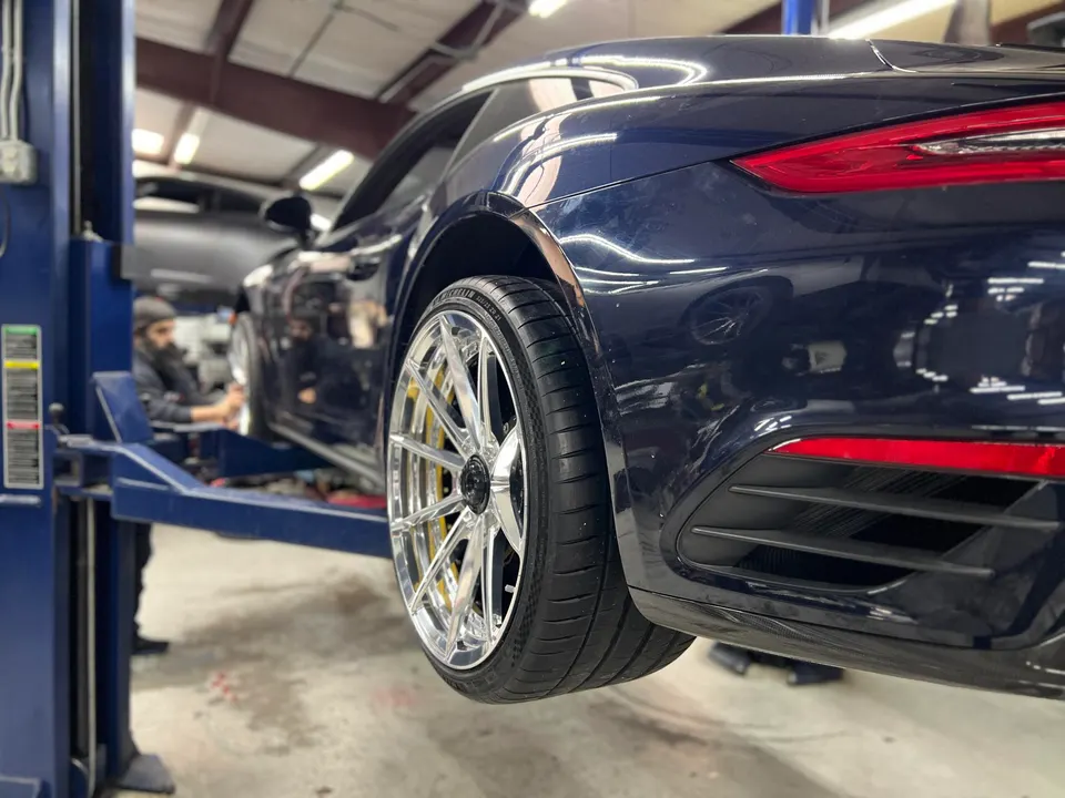 A dark blue Porsche 911 on a lift in a shop, showcasing custom chrome wheels and tires with a mechanic in the background.