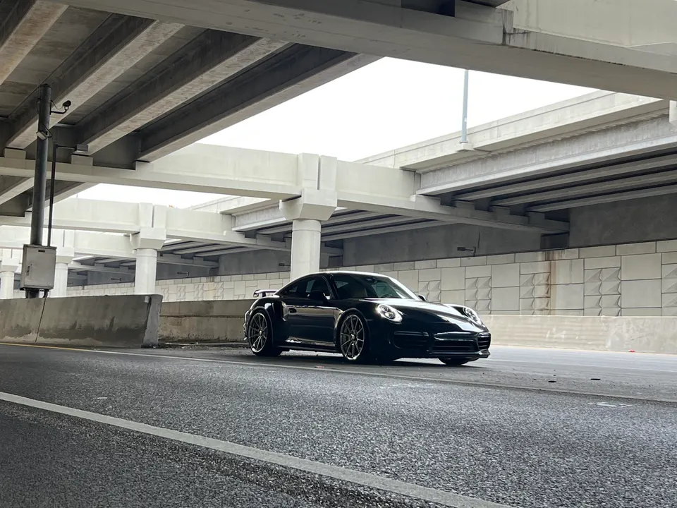 Black Porsche 911 sports car parked under a concrete overpass on an urban road