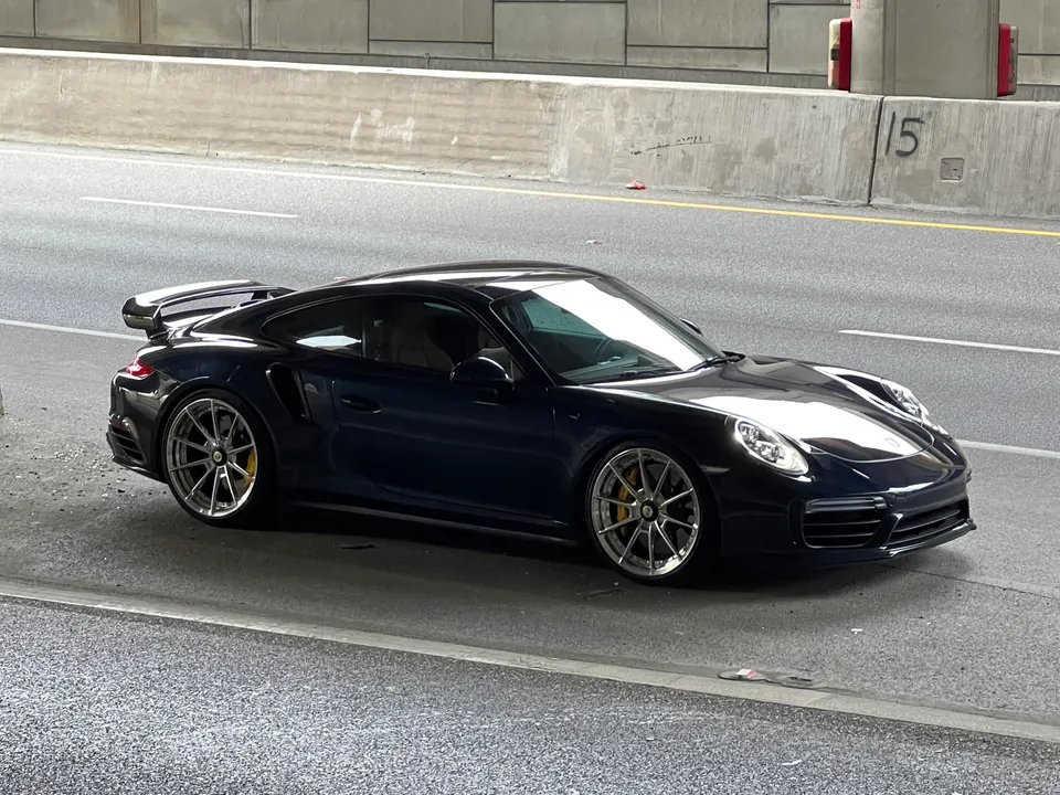Dark blue Porsche 911 Turbo S parked on the side of a road under an overpass, showcasing its sleek design and aftermarket wheels.