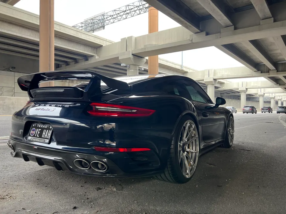 Black Porsche 911 Turbo S with aftermarket wheels, large rear wing, and carbon fiber diffuser parked under an overpass.