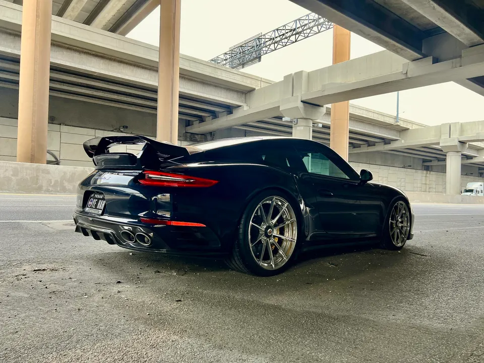 A black Porsche 911 with a large rear wing and custom wheels parked under an overpass.