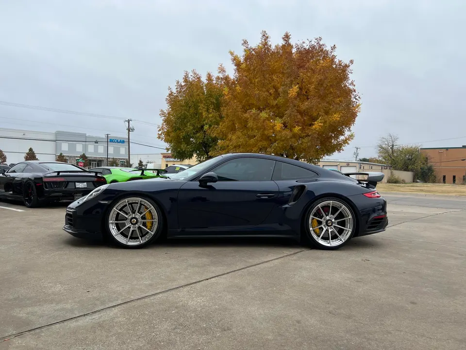 Dark blue Porsche 911 Turbo S with aftermarket multi-spoke wheels and yellow brake calipers parked outdoors.