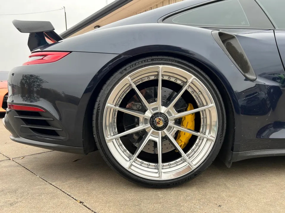 A close-up of the rear wheel of a dark blue Porsche 911, showcasing custom silver wheels with yellow brake calipers and a carbon fiber wing.