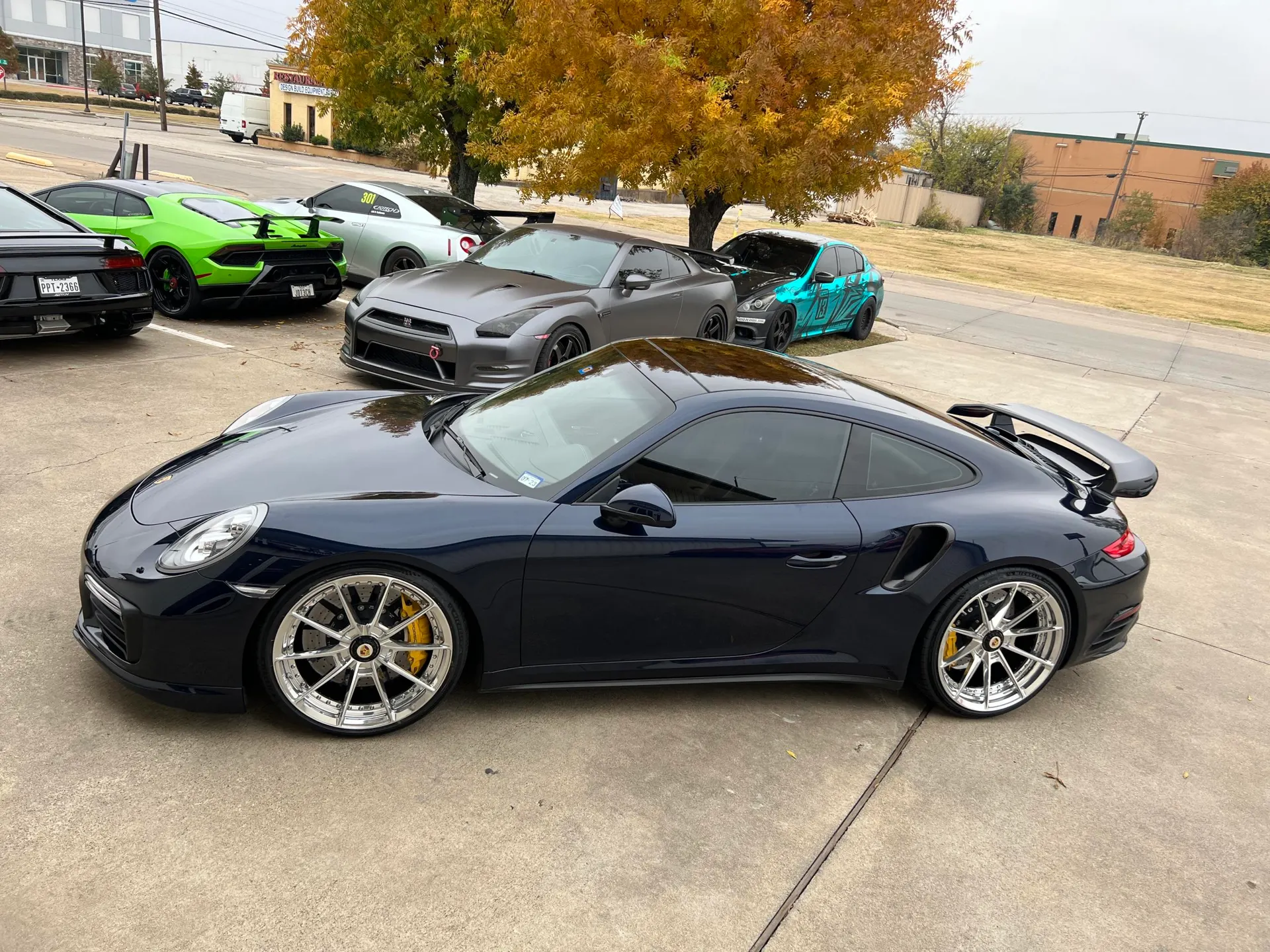 A dark blue Porsche 911 Turbo S with custom wheels parked alongside a green Lamborghini Huracan and several Nissan GT-Rs in a parking lot.