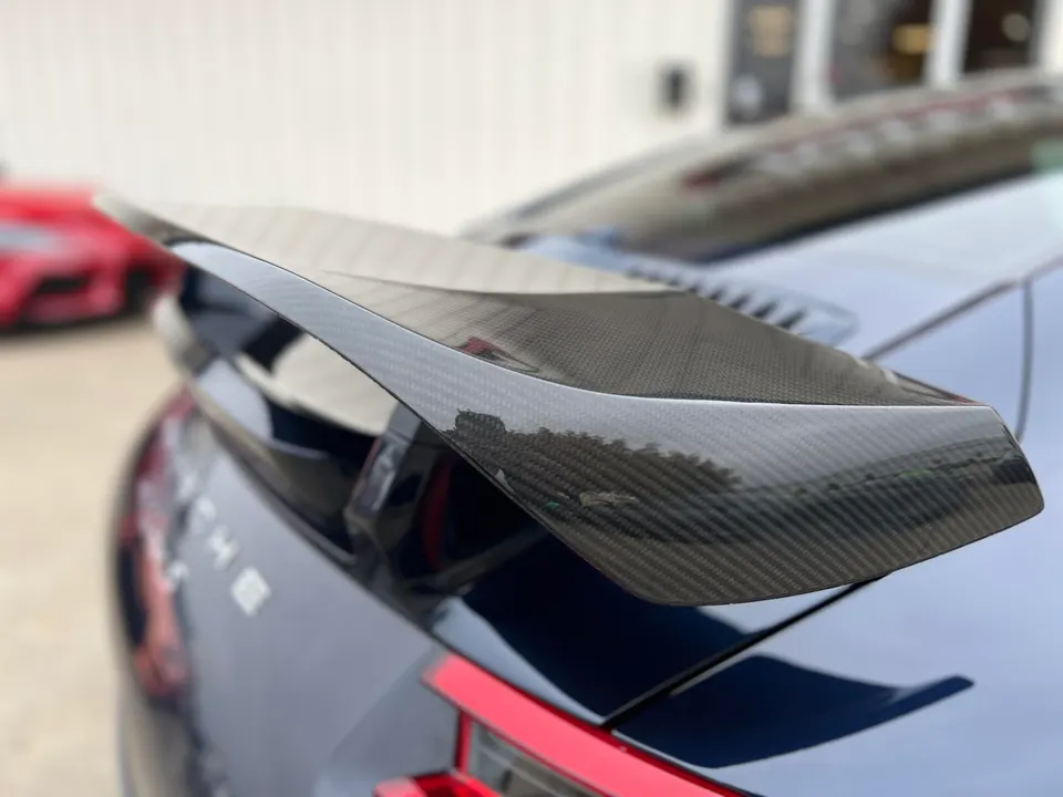 A close-up of a carbon fiber rear wing on a dark blue Porsche, with a red car blurred in the background.